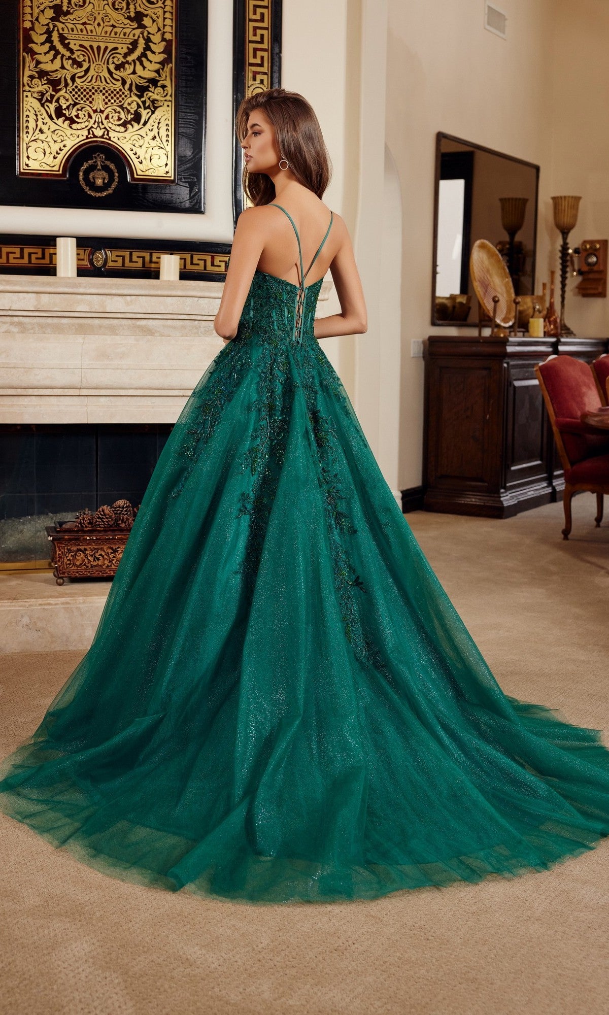 A woman wearing the Nox Anabel H1883 long prom dress, featuring a green lace corset bodice, stands with her back to the camera in an elegantly decorated living room.
